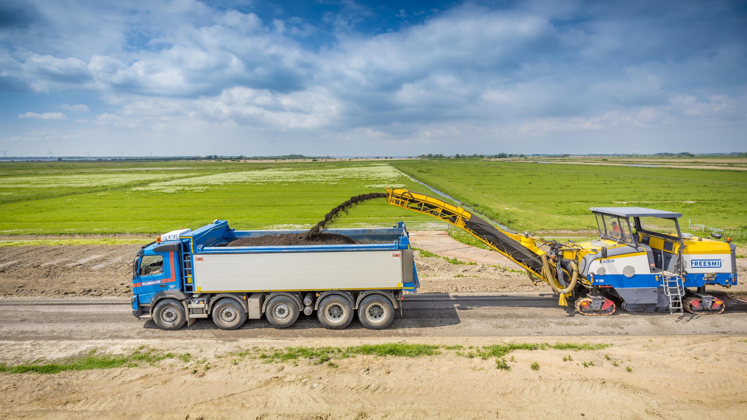 Blauwe Ploegam vrachtwagen krijgt zand aangevoerd met groen weiland op de achtergrond
