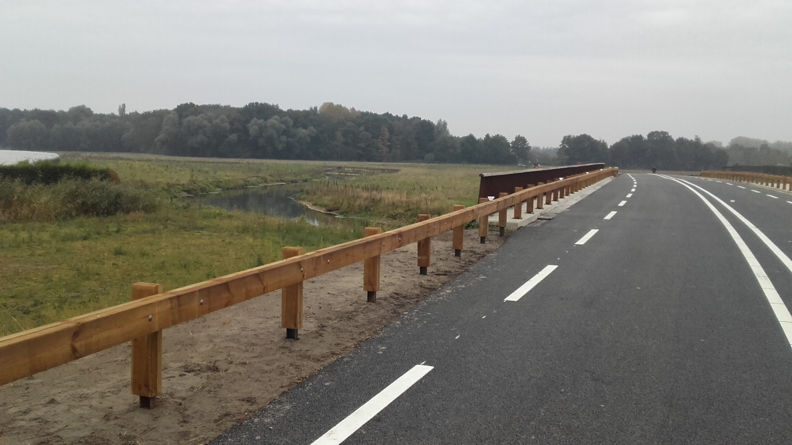 Weg met houten vangrail naast een natuurgebied met een beekje en gras