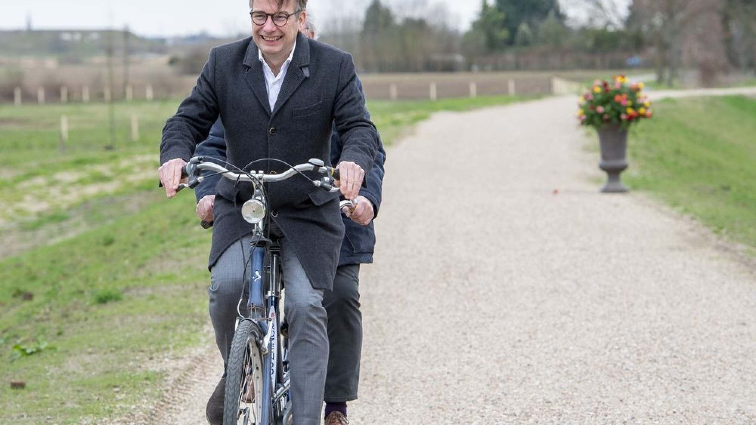 Twee personen fietsen op een tandem over een weg met bloempot in de achtergrond