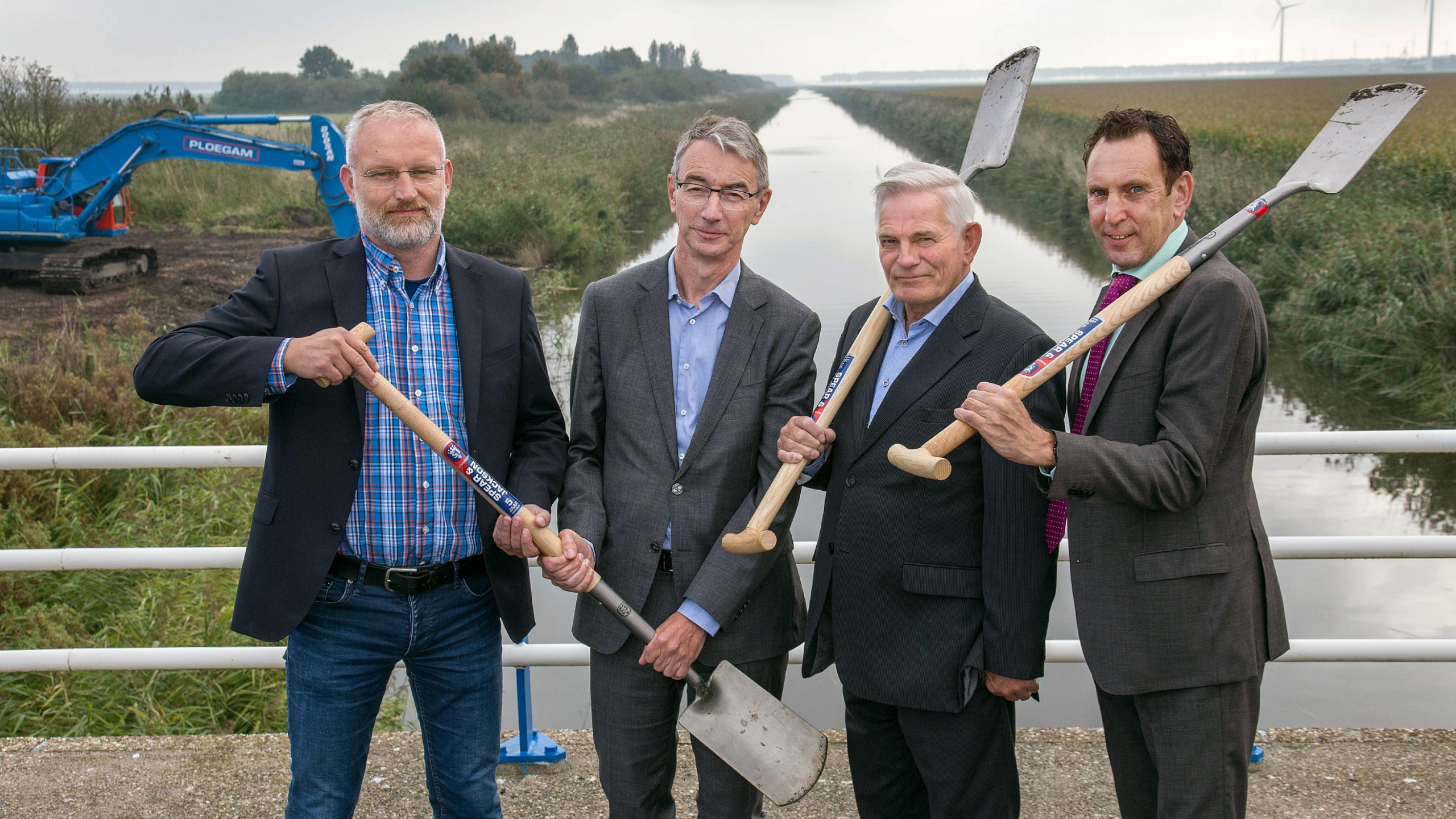 Vier mannen in pak met scheppen in de hand en Ploegam kraan op de achtergrond