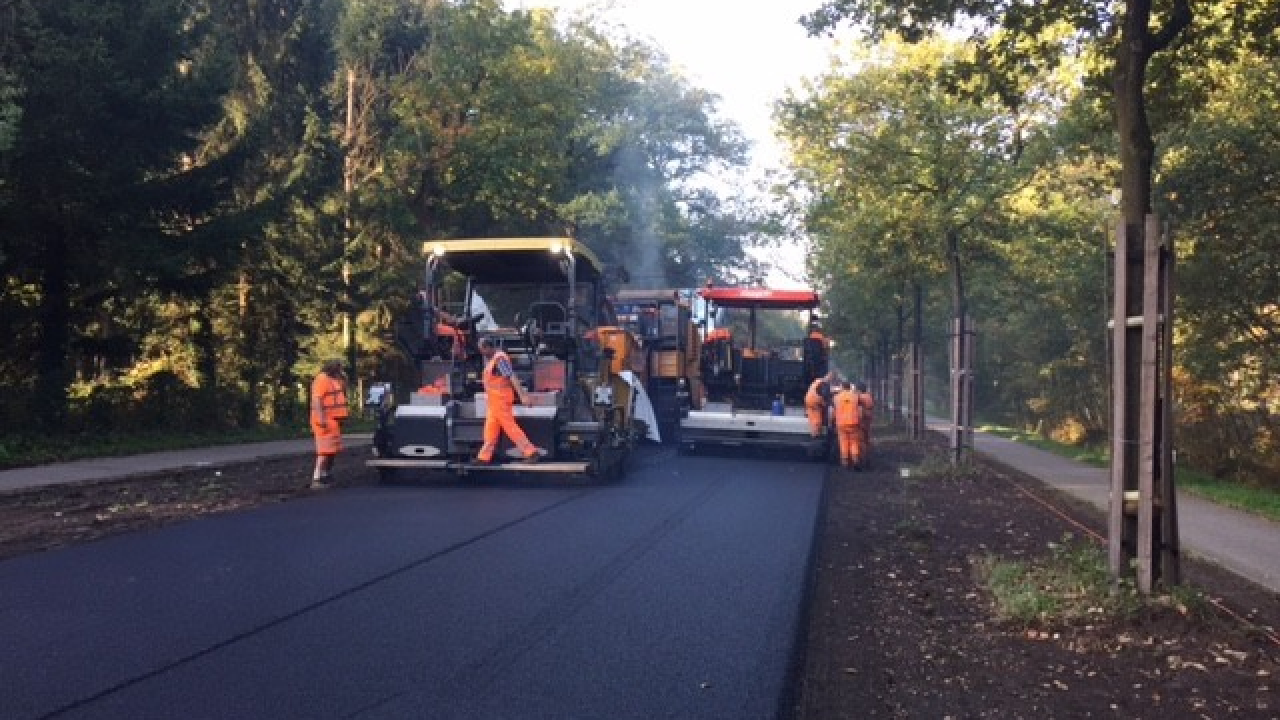 Mannen in oranje veiligheidskleding lopen naast machines op nieuw geasfalteerde weg