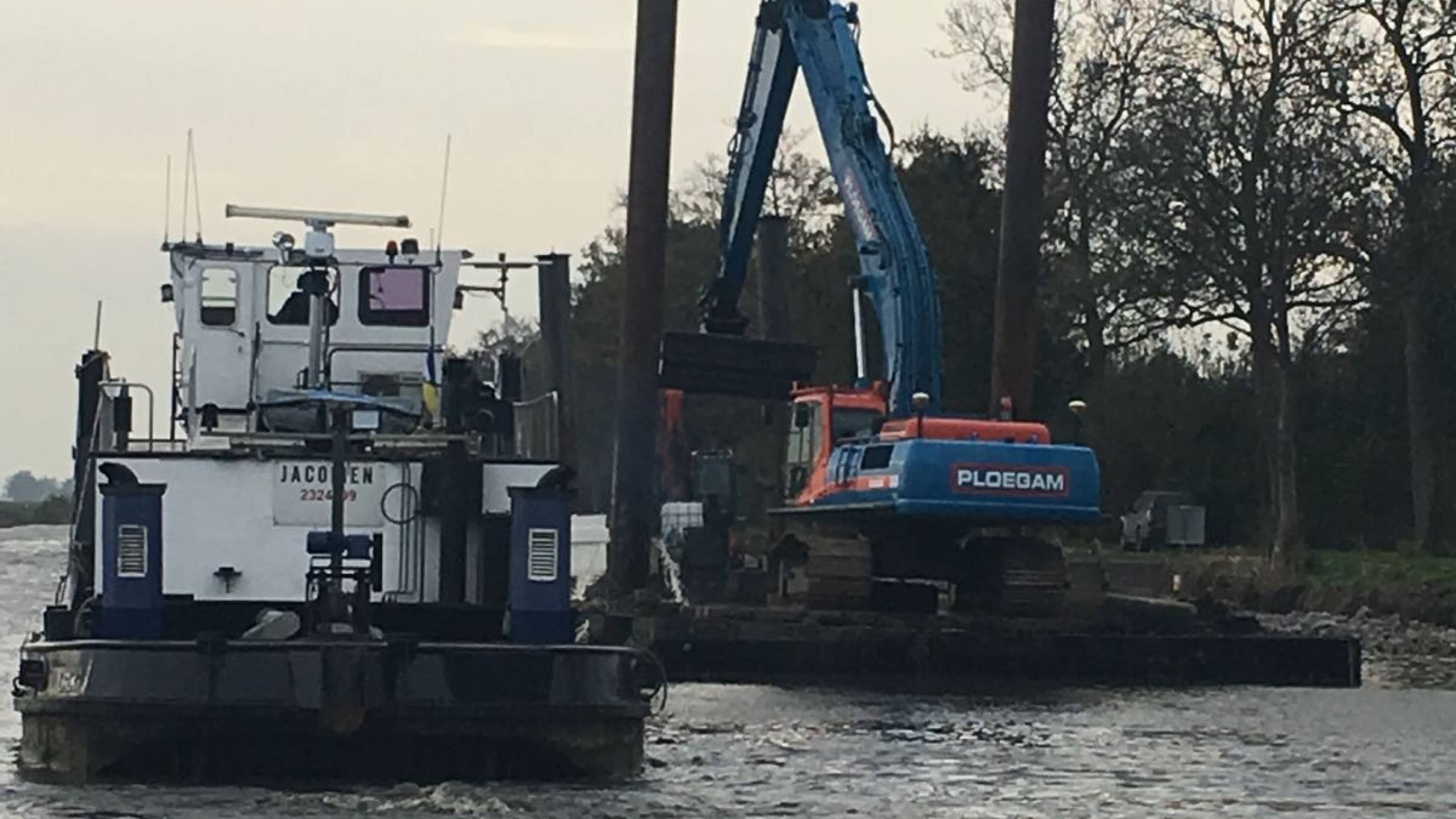 Blauwe Ploegam kraan werkt vanaf ponton op het water met een witte werkboot ernaast