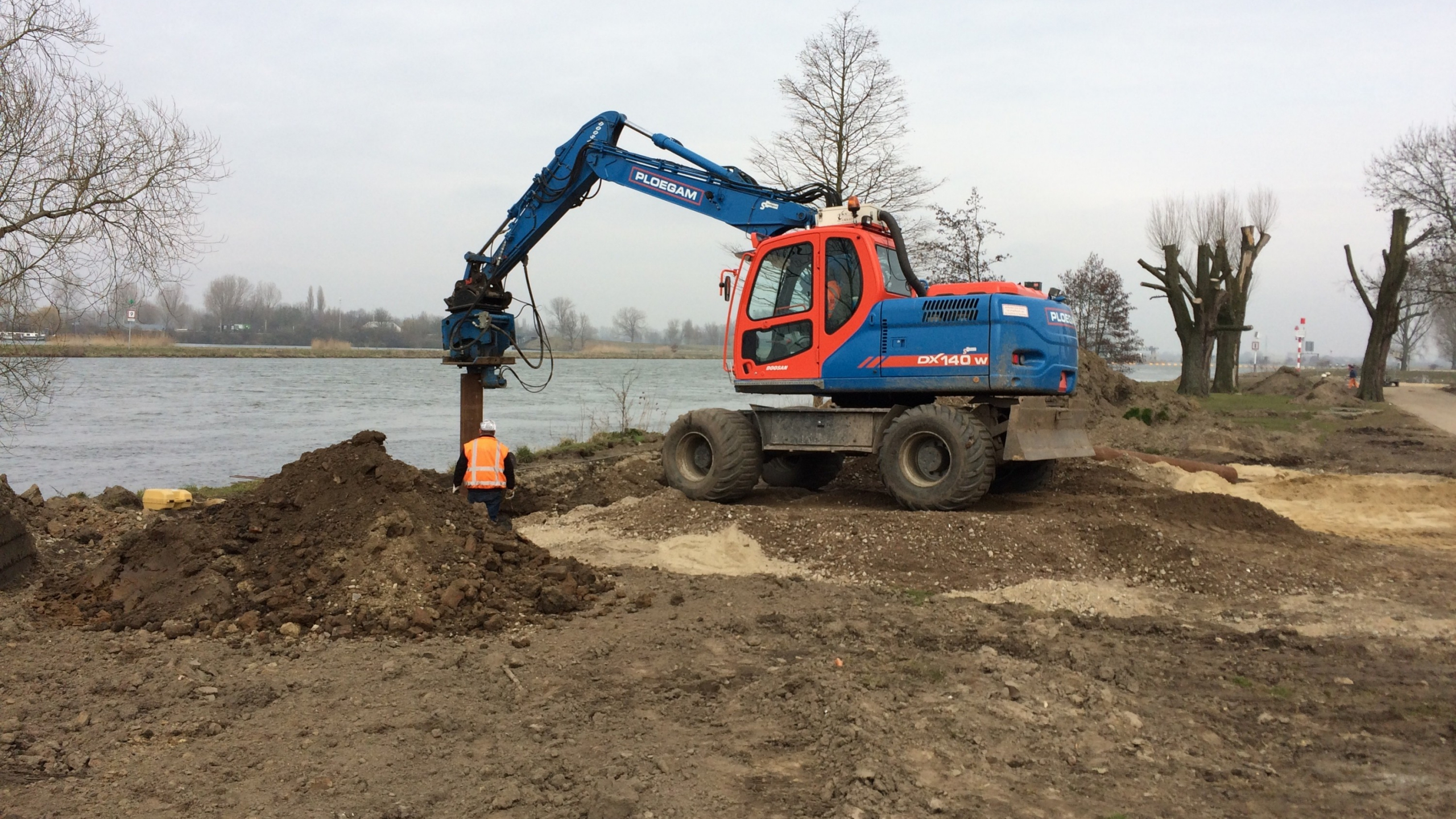 Blauwe Ploegam graafmachine in het zand naast een rivier in Roermond
