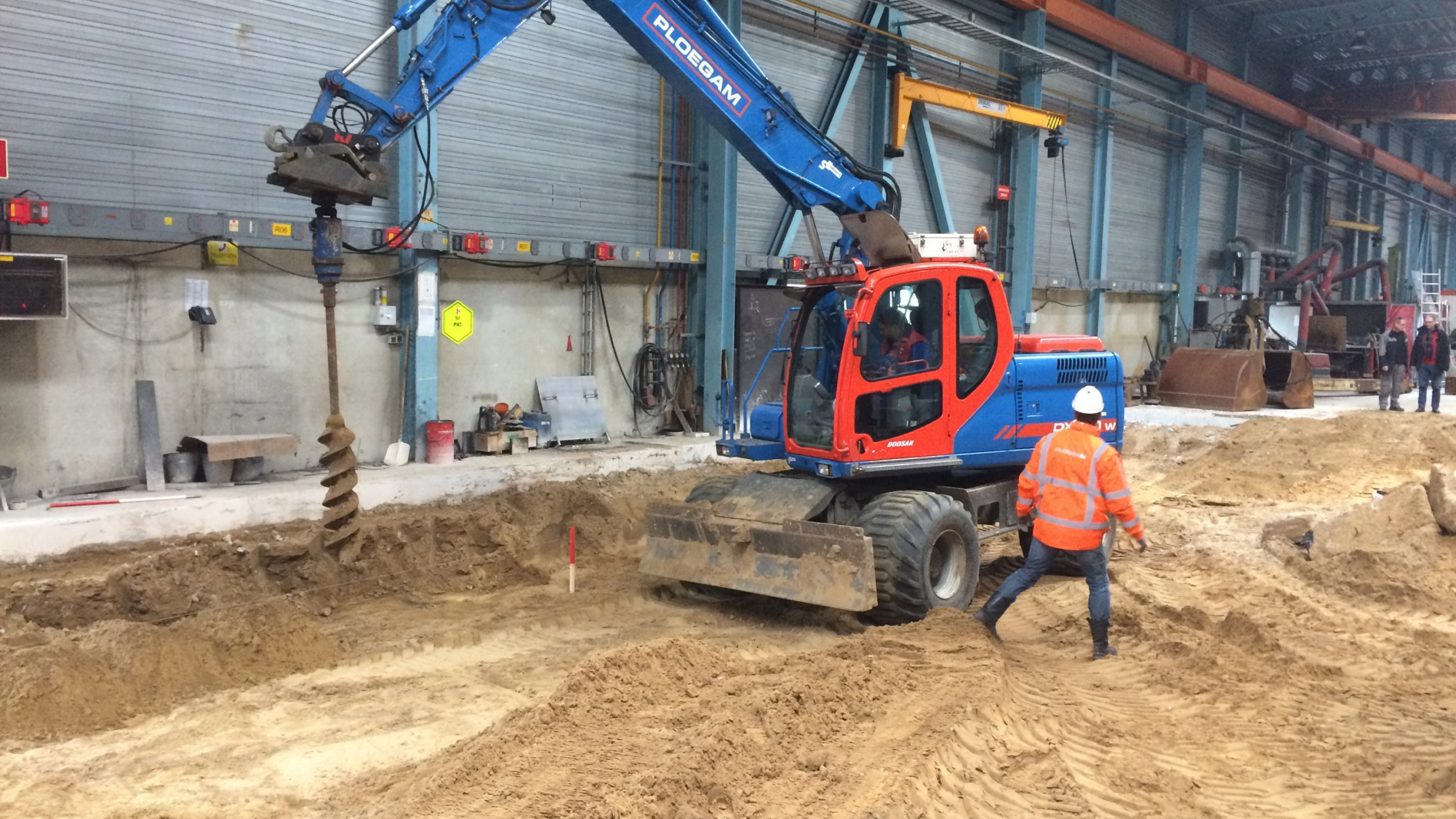 Man met veiligheidsjas en helm loopt langs blauwe kraan in het zand