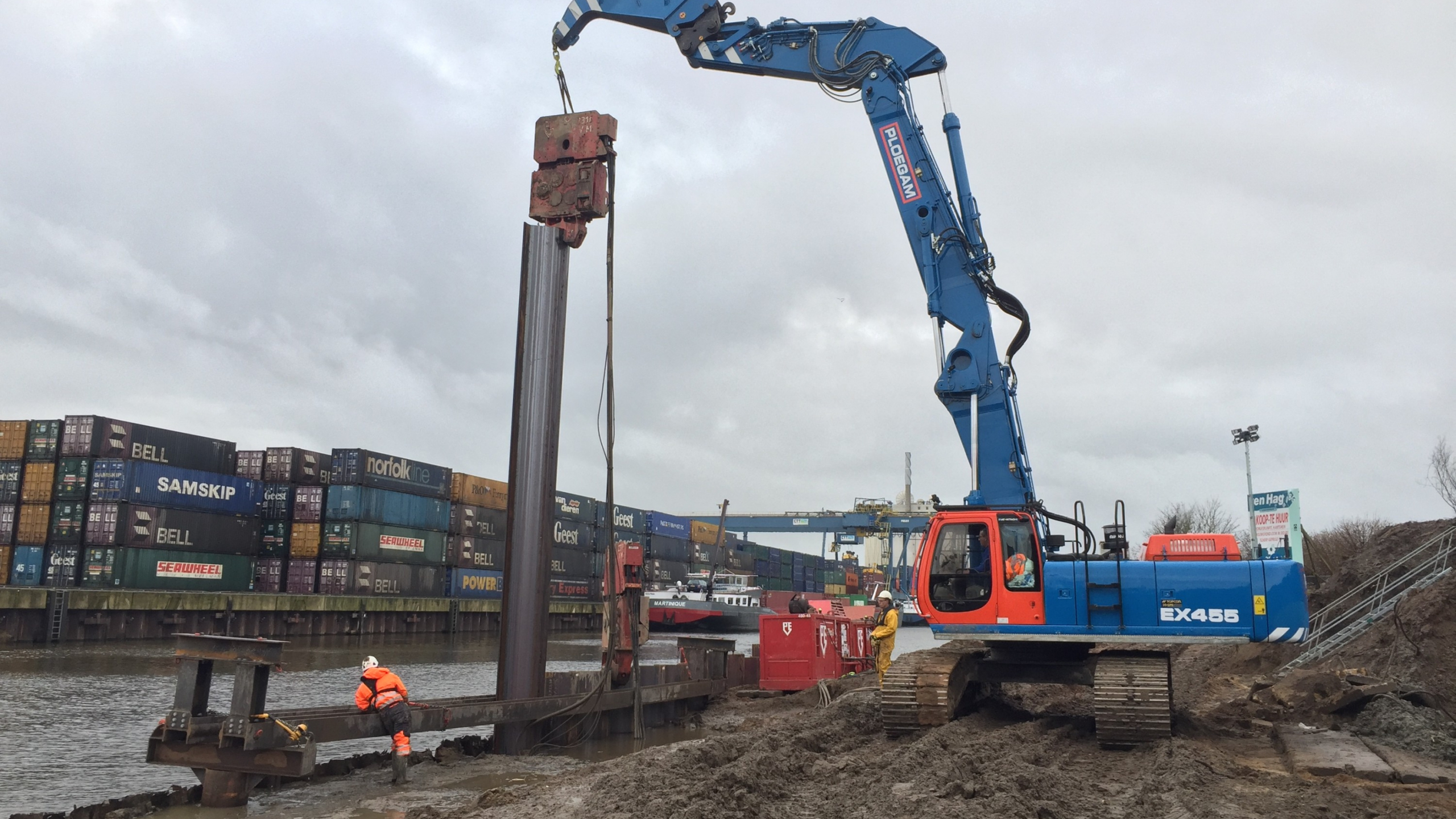 Blauwe Ploegam kraan plaatst damwand met containerschip op de achtergrond