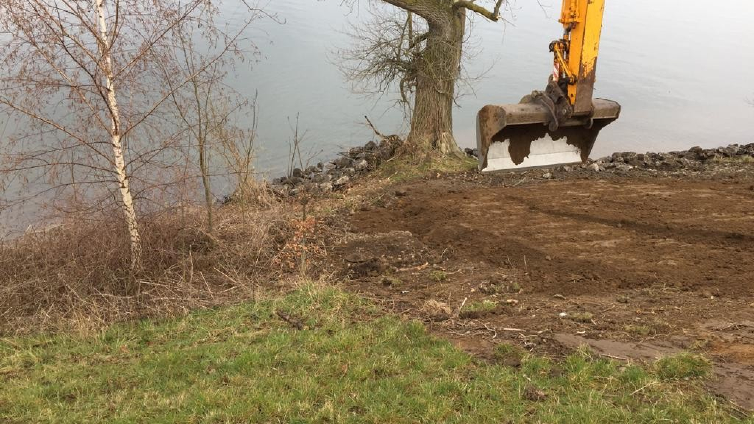 Bak van gele kraan schept zand naast een rivier