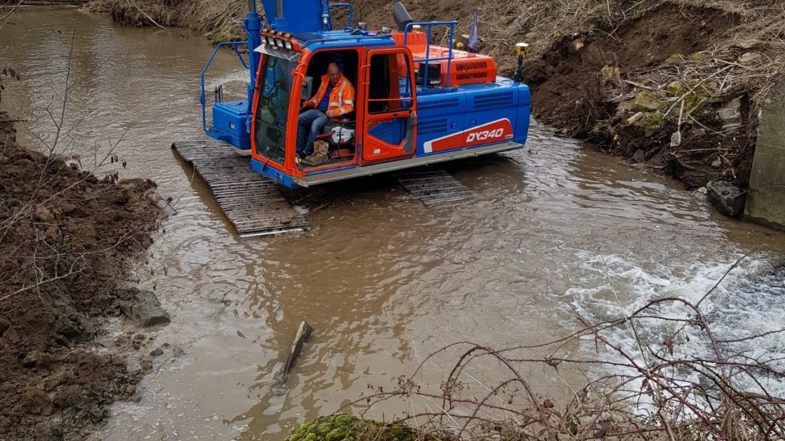 Man met veiligheidsjas zit in een blauwe Ploegam kraan die in bruin water staat