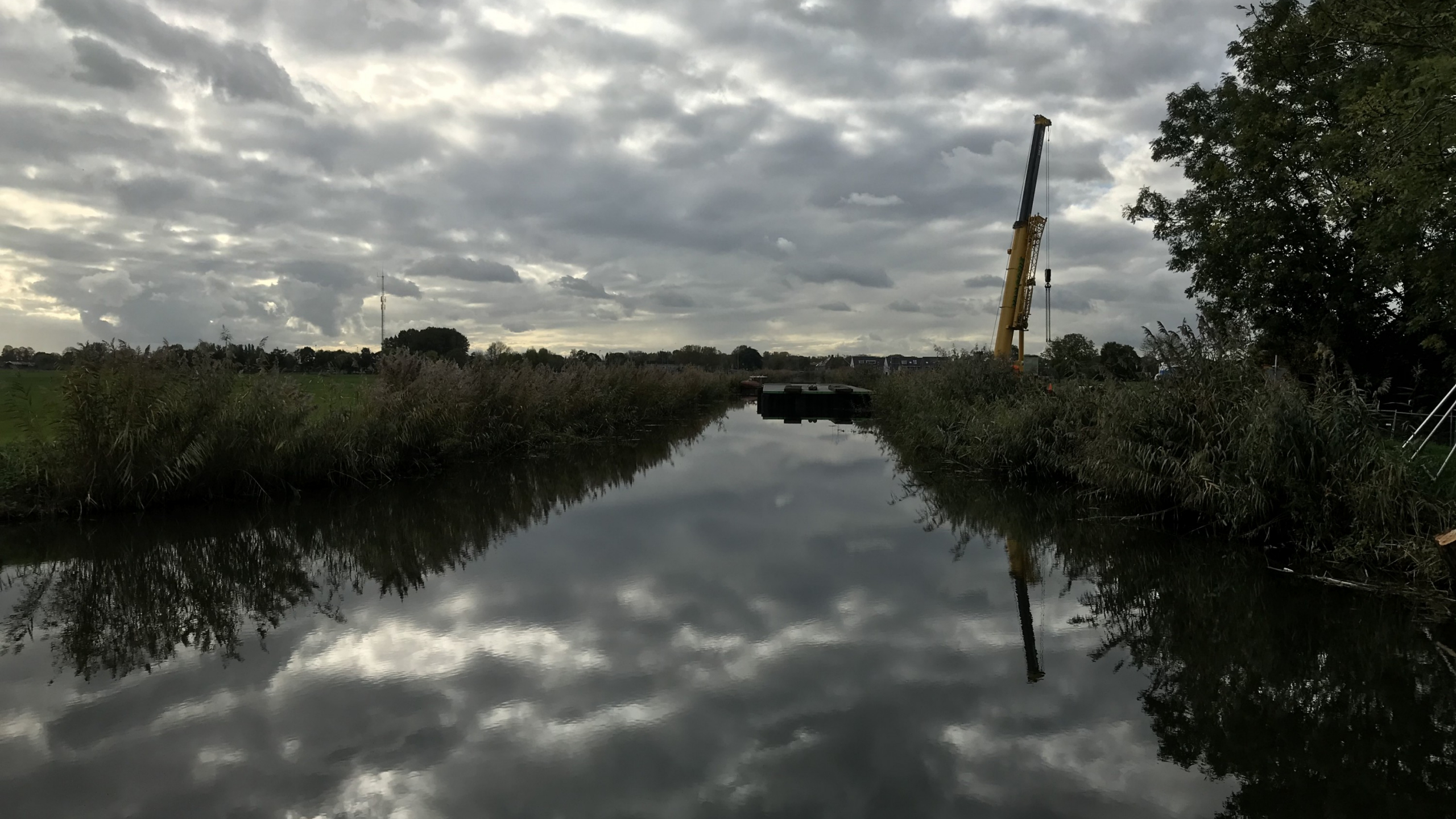 Natuurfoto met water en struiken en een gele kraan op de achtergrond
