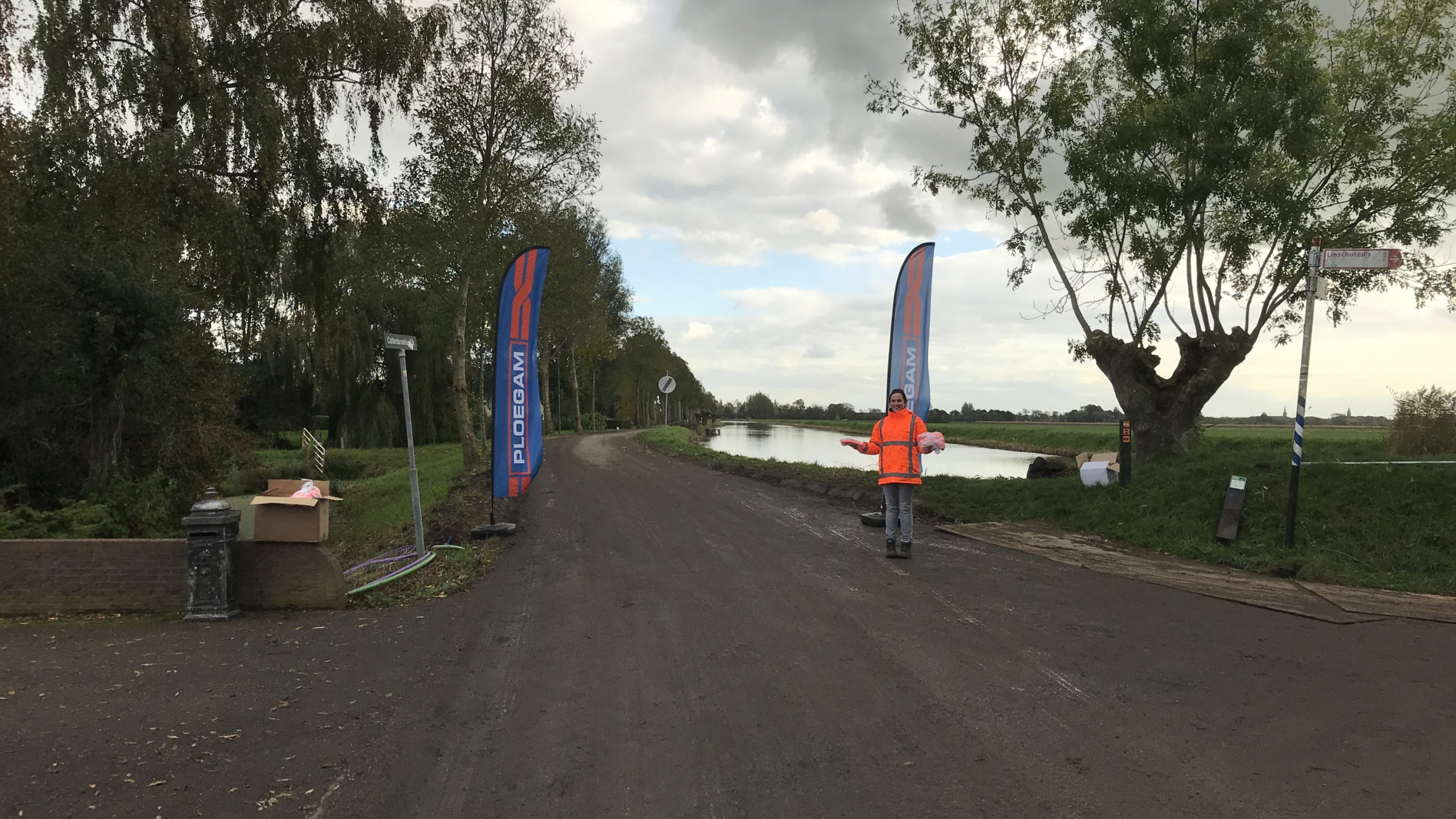 Vrouw met oranje veiligheidsjas poseert voor twee Ploegam vlaggen op een weg naast een rivier