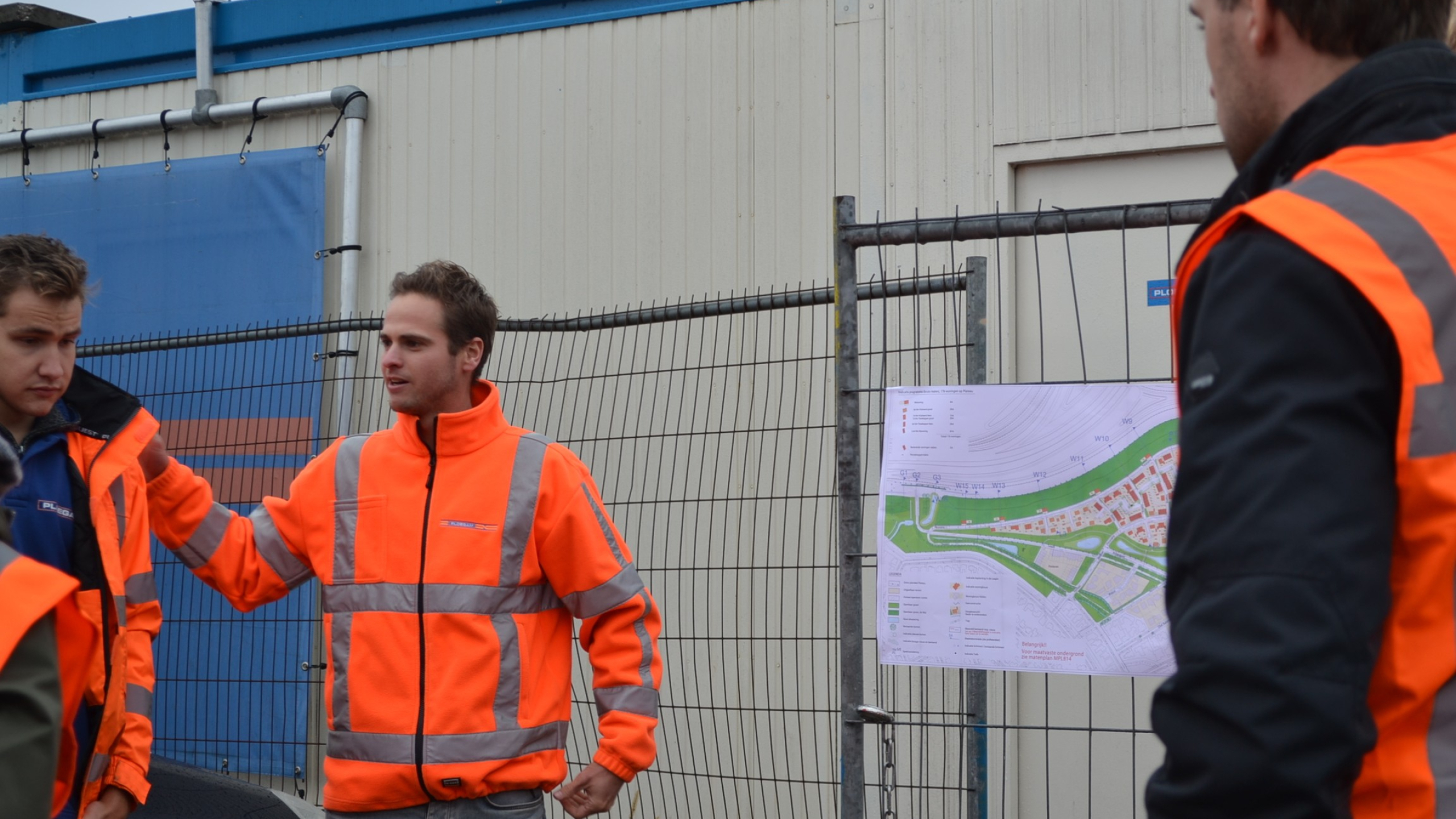 Medewerkers van Ploegam in oranje jassen in overleg met een plattegrond van de werkzaamheden op een bouwhek