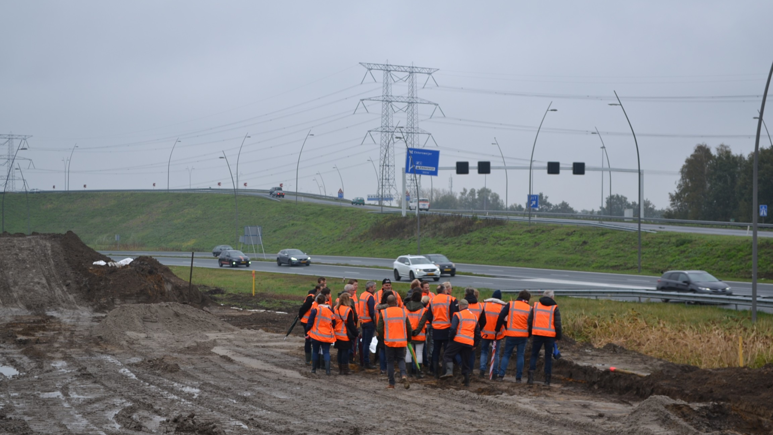 Groep personen in oranje veiligheidshesjes op bouwplaats in het zand naast een snelweg