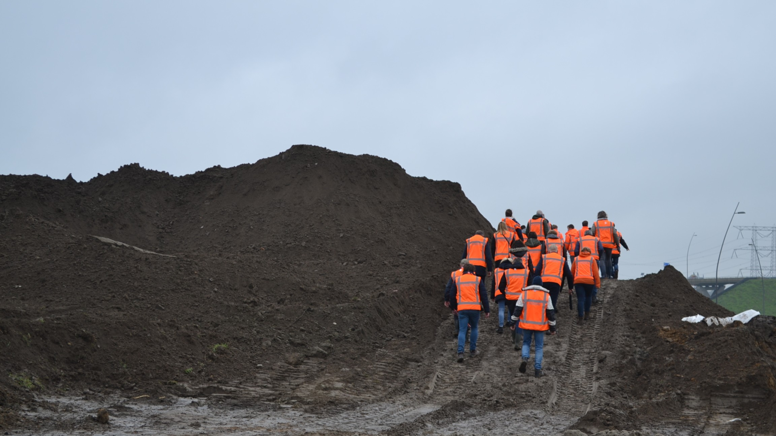 Groep mensen in oranje veiligheidsvesten loopt over een heuvel van zwart zand op een bouwplaats