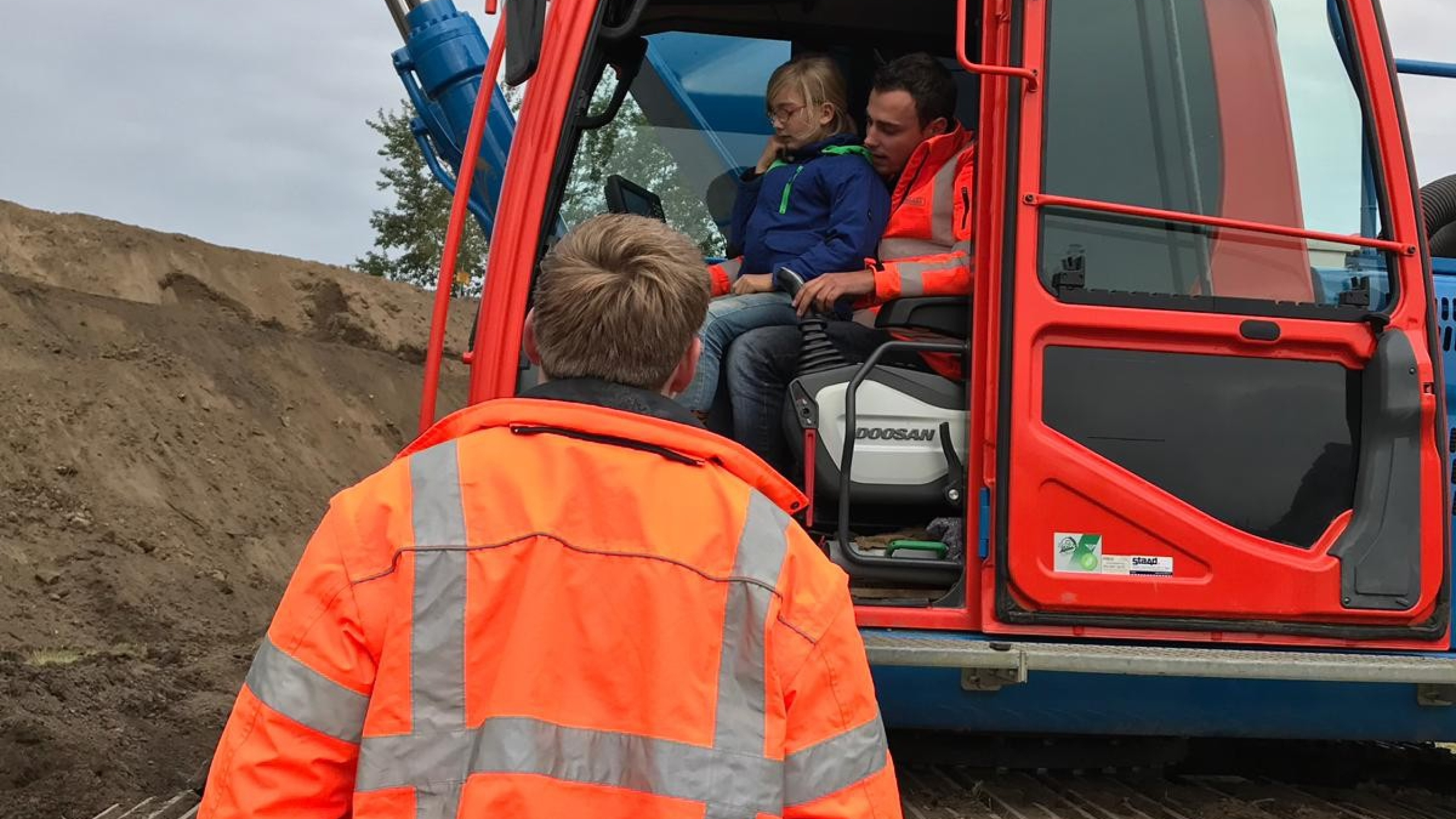 Man met oranje veiligheidsjas kijkt naar man met kind in de cabine van een kraan