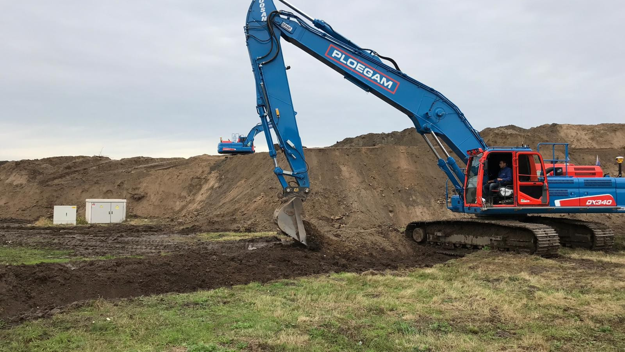 Blauwe Ploegam rupskraan schept zand op een bouwplaats met een andere kraan op de achtergrond