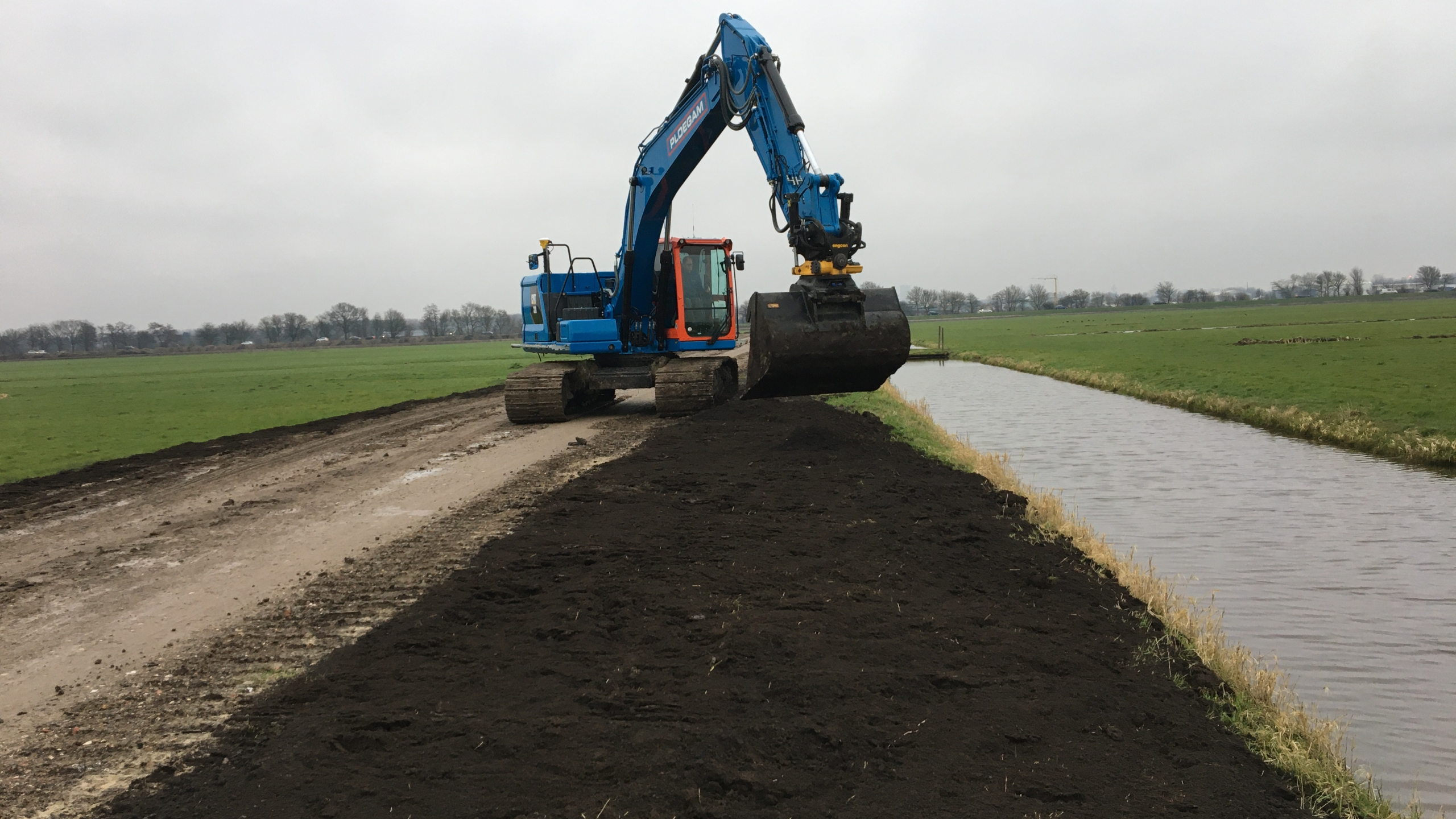 Blauwe kraan rijdt over zandweg naast een rivier