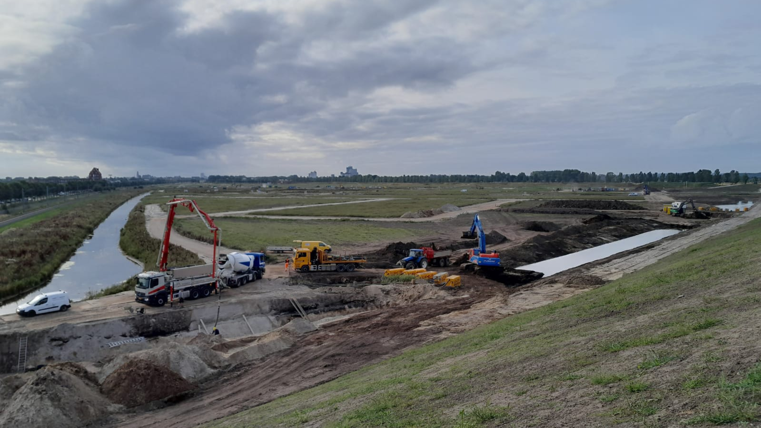 Een kraan, vrachtwagens, tractor en busje op het zand naast een beekje
