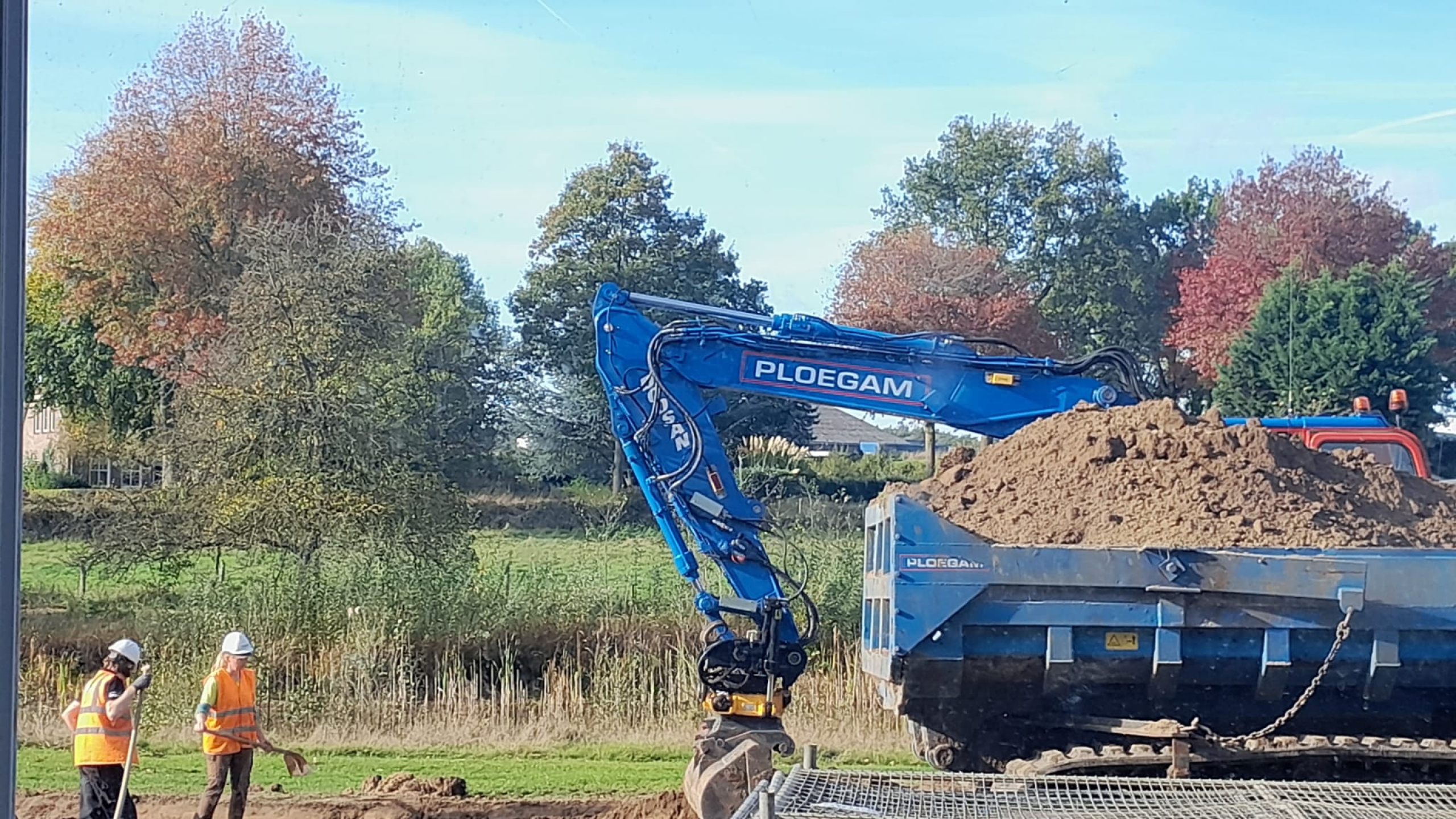 Blauwe Ploegam kraan schept zand en twee Ploegam medewerkers kijken toe