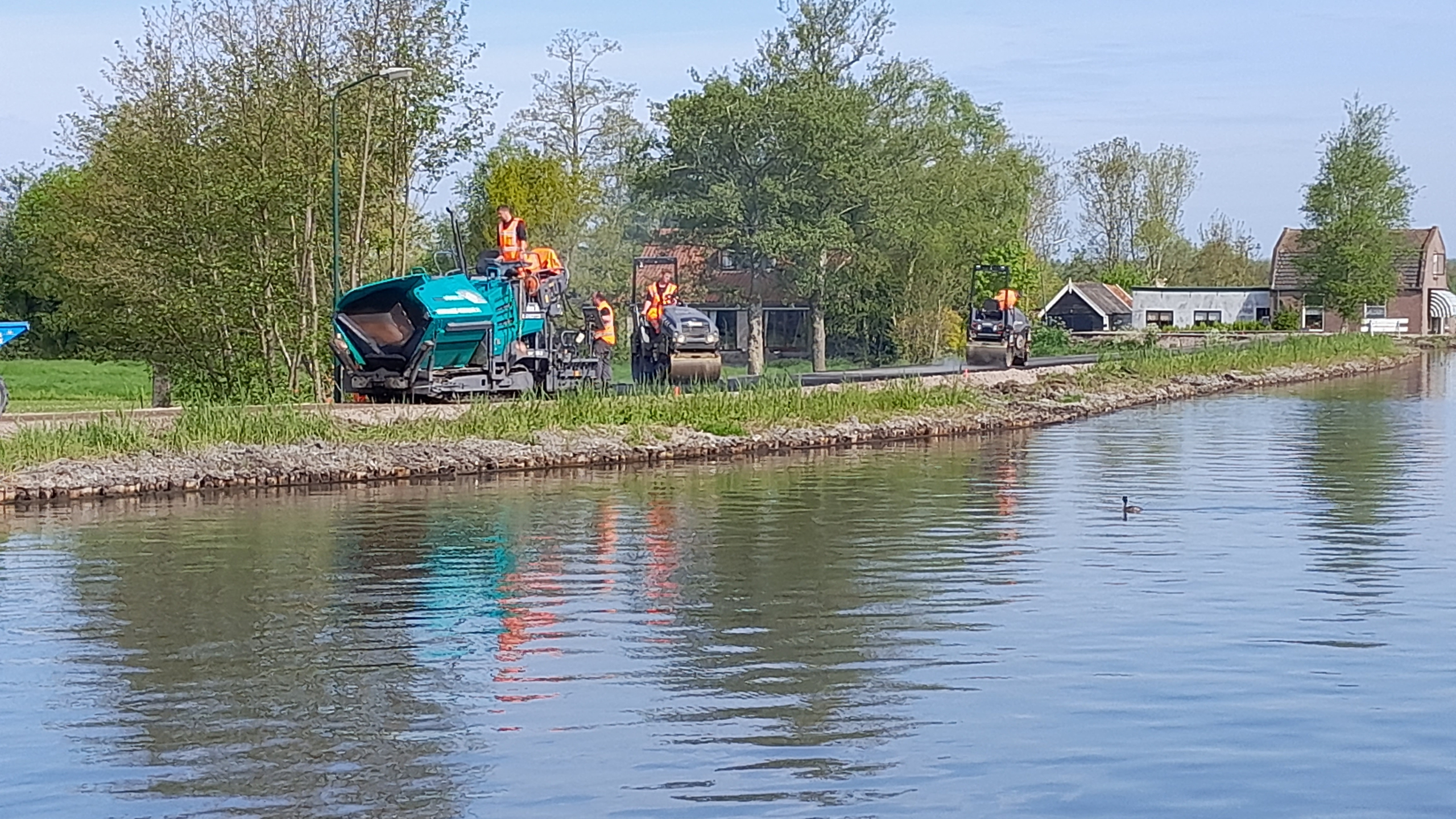 Werkzaamheden naast rivier met huizen op de achtergrond
