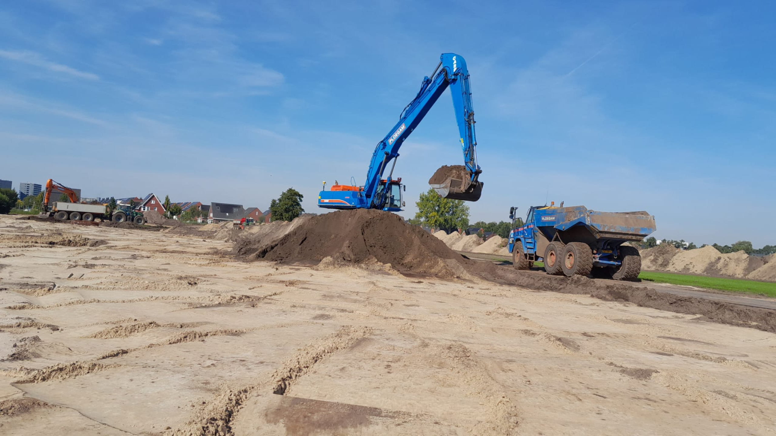 Blauwe Ploegam kraan en kieper in het zand met huizen op de achtergrond