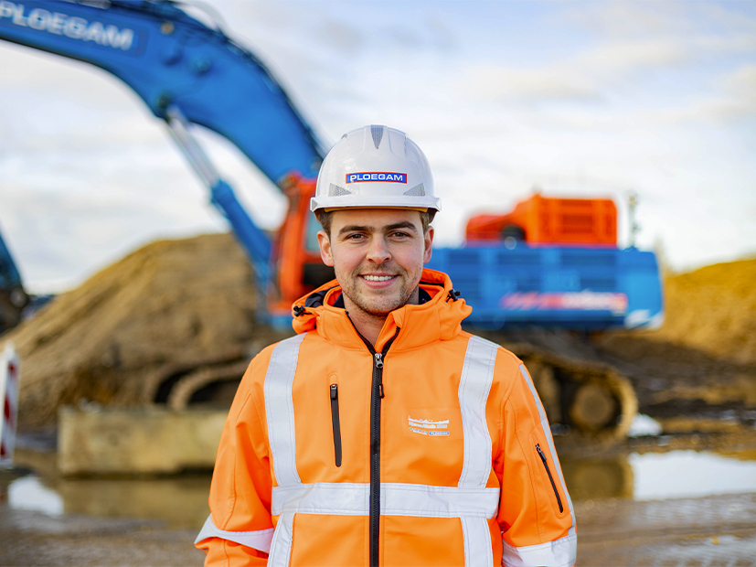 Man met oranje jas en witte helm met een blauwe kraan op de achtergrond