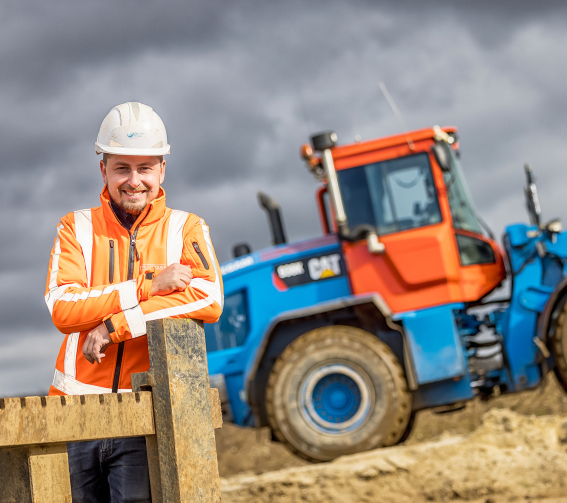 Man met veiligheidsjas en helm op bouwplaats met blauwe shovel op de achtergrond