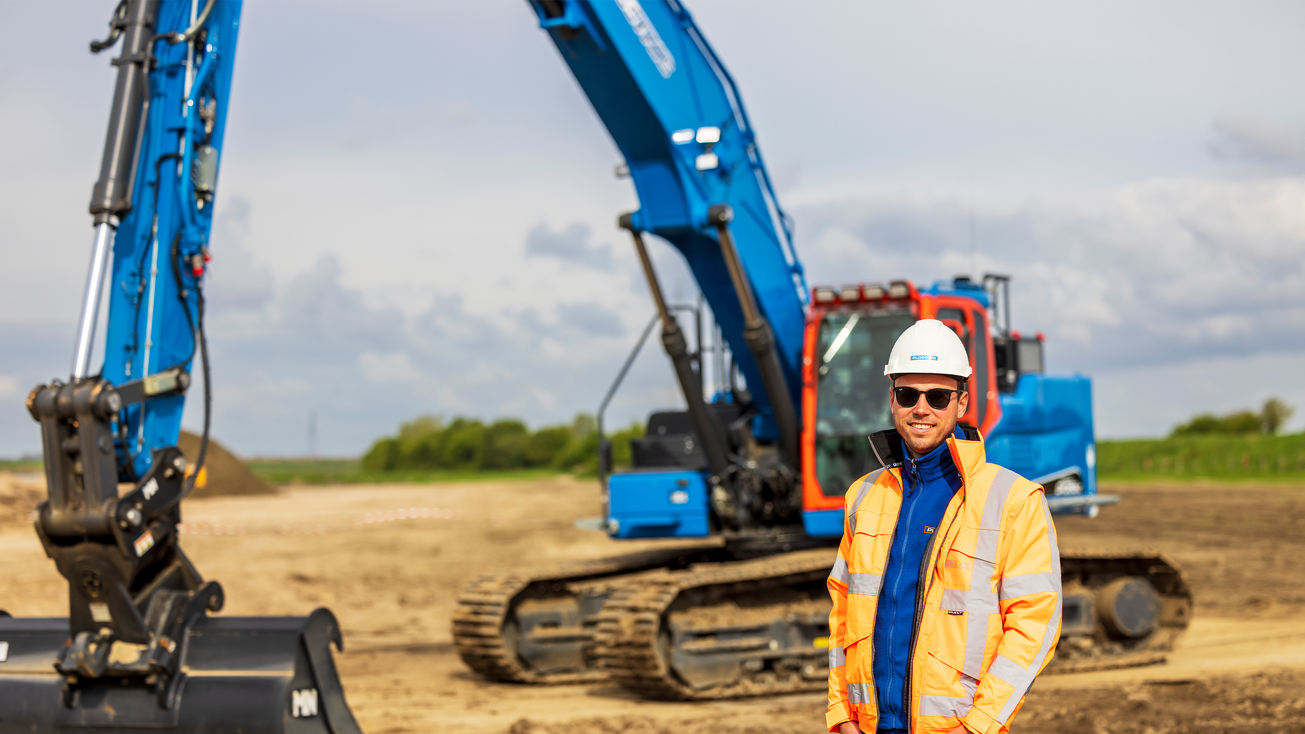 Ploegam medewerker met oranje jas en helm met een blauwe kraan en zand op de achtergrond