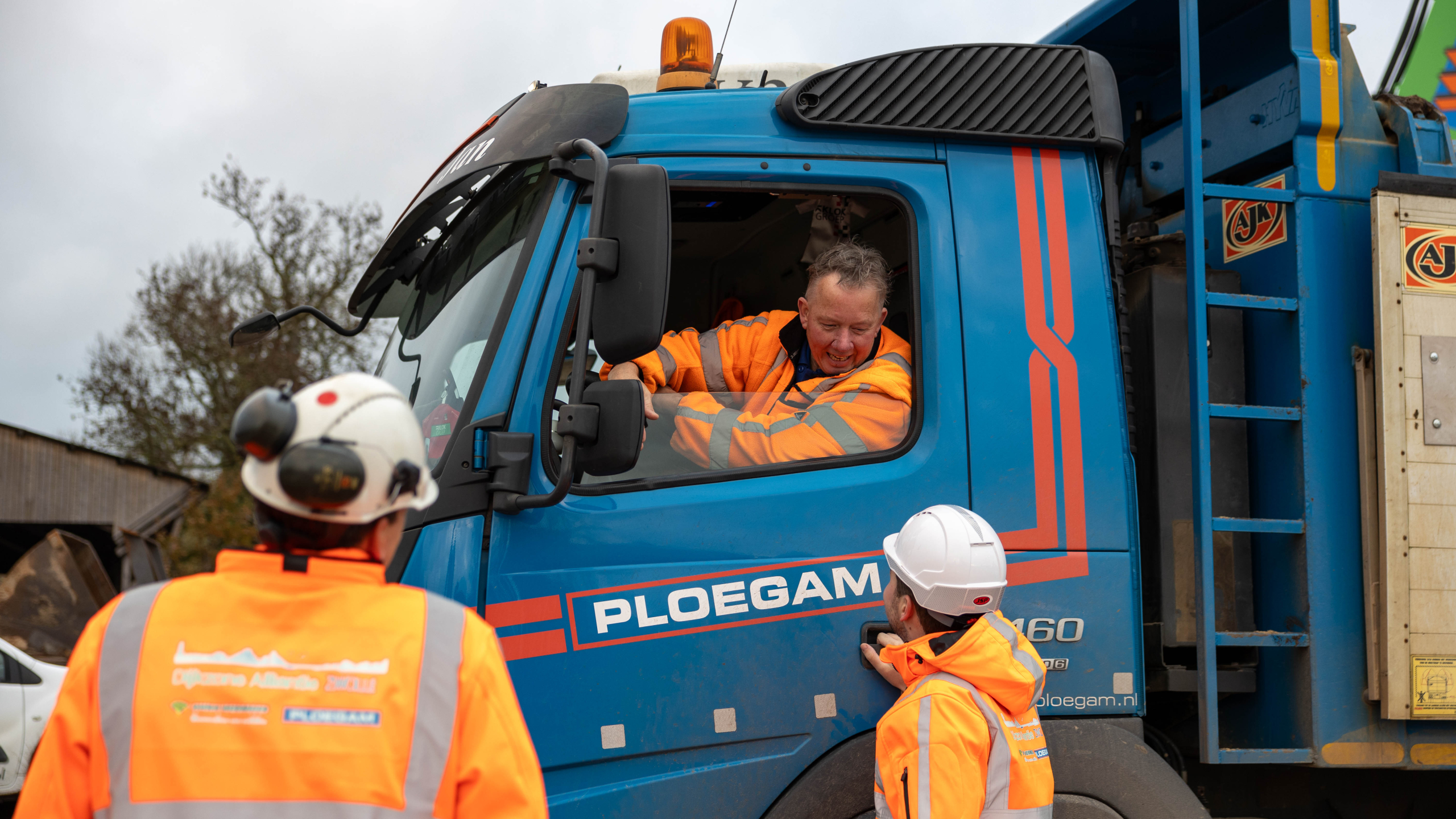 Twee mannen met oranje jassen en helmen in overleg met chauffeur van Ploegam vrachtwagen