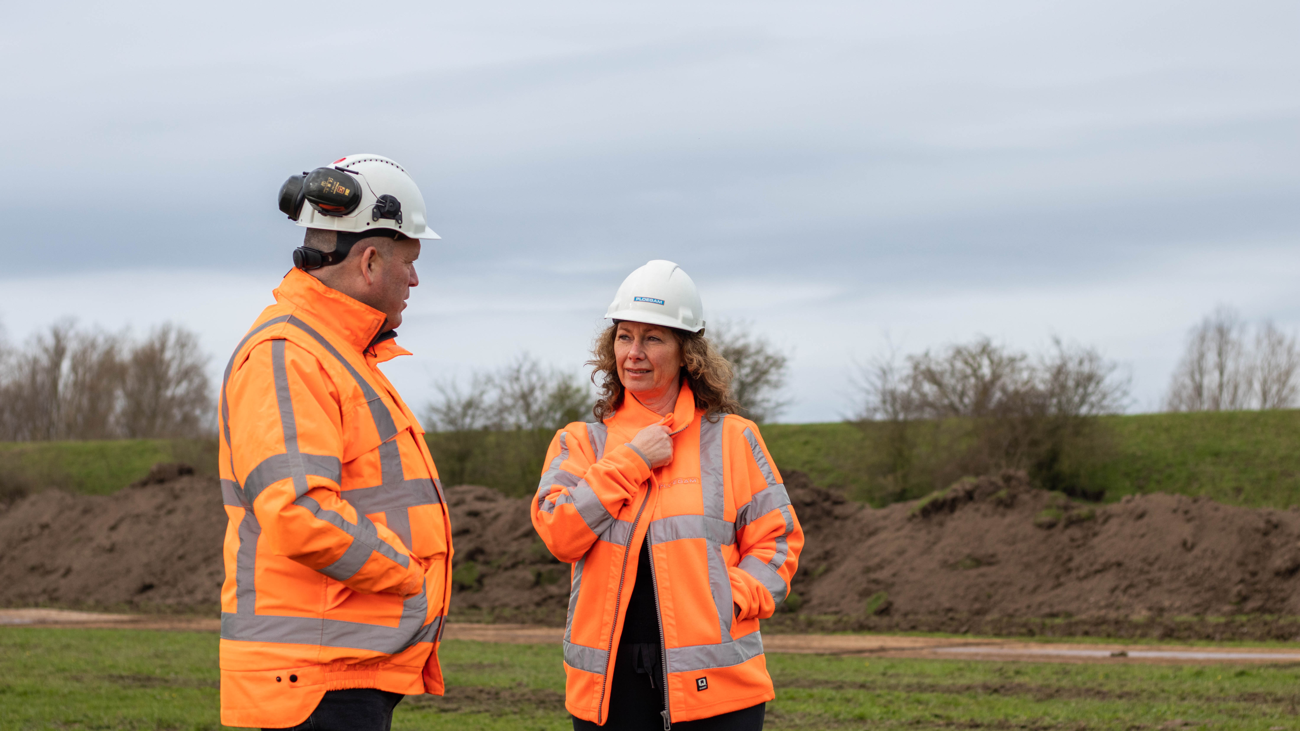 Man en vrouw met oranje veiligheidsjassen en helmen in overleg buiten