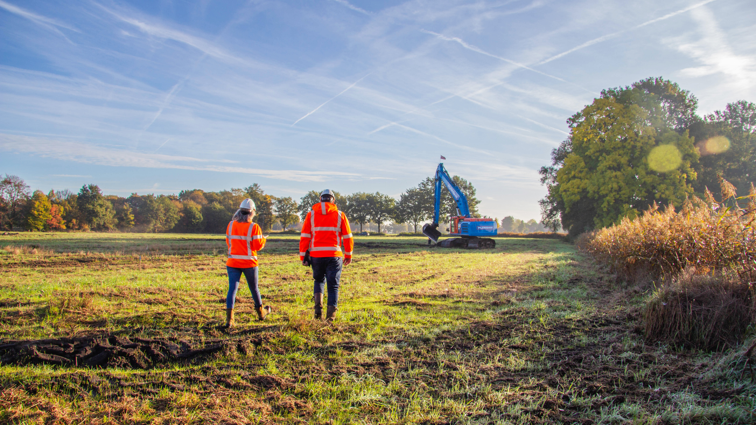 Vrouw en man met oranje veiligheidsjassen lopen over het gras met een blauwe kraan op de achtergrond
