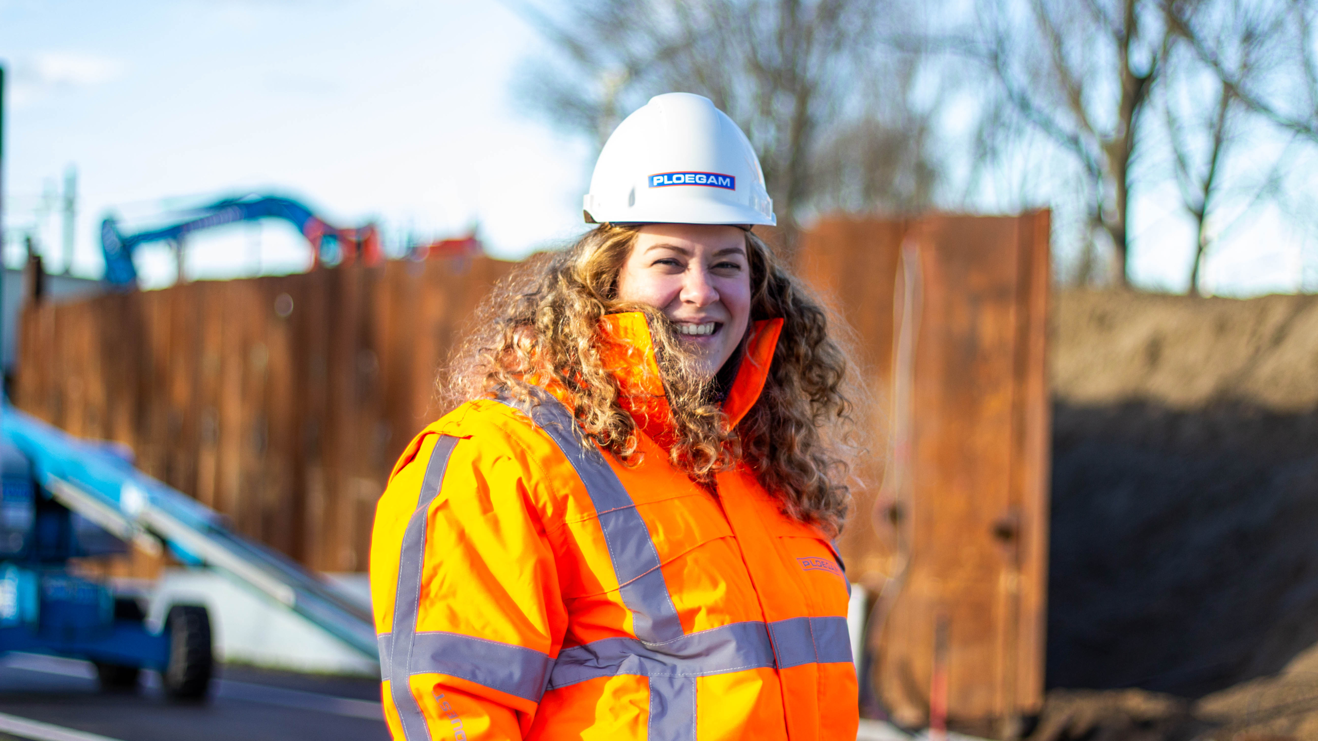 Ploegam medewerkster met oranje jas en witte helm met damwanden op de achtergrond