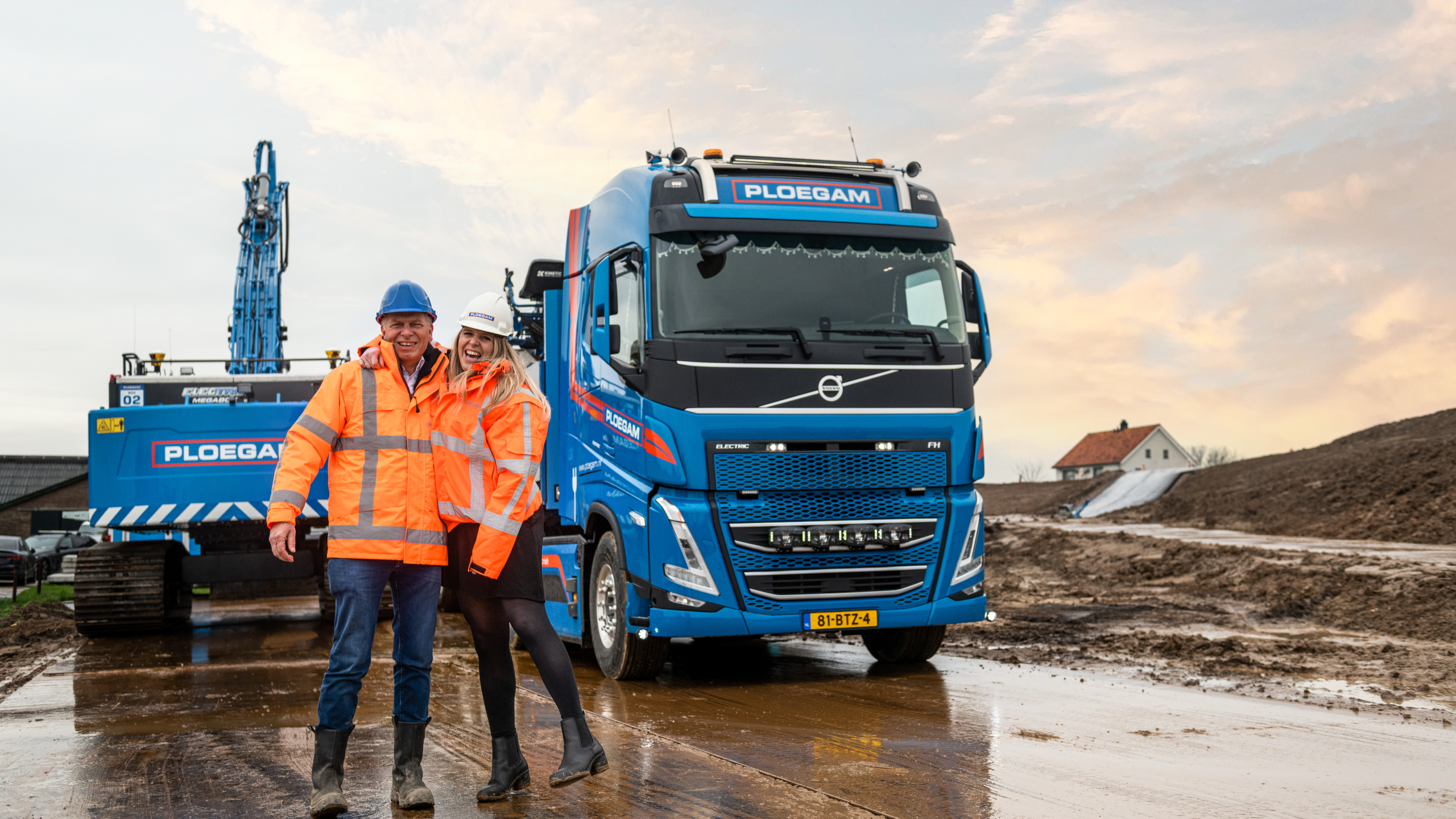 Lachende man en vrouw met oranje veiligheidsjassen en helmen voor een blauwe kraan en vrachtwagen