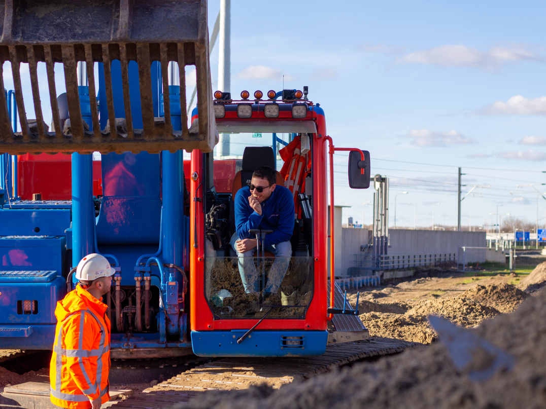 Man met oranje jas en helm in overleg met kraanmachinist in een blauwe kraan