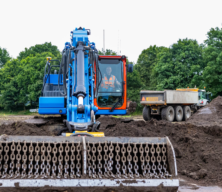 Man met veiligheidshesje zit in een blauwe kraan op bouwplaats met zand