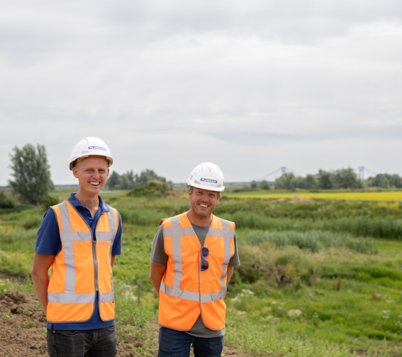 Man met oranje jas en witte Ploegam helm met een blauwe kraan op de achtergrond