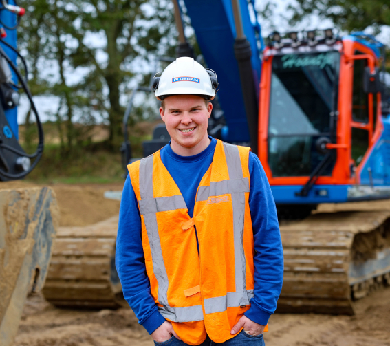 Man met oranje jas en witte Ploegam helm met een blauwe kraan op de achtergrond