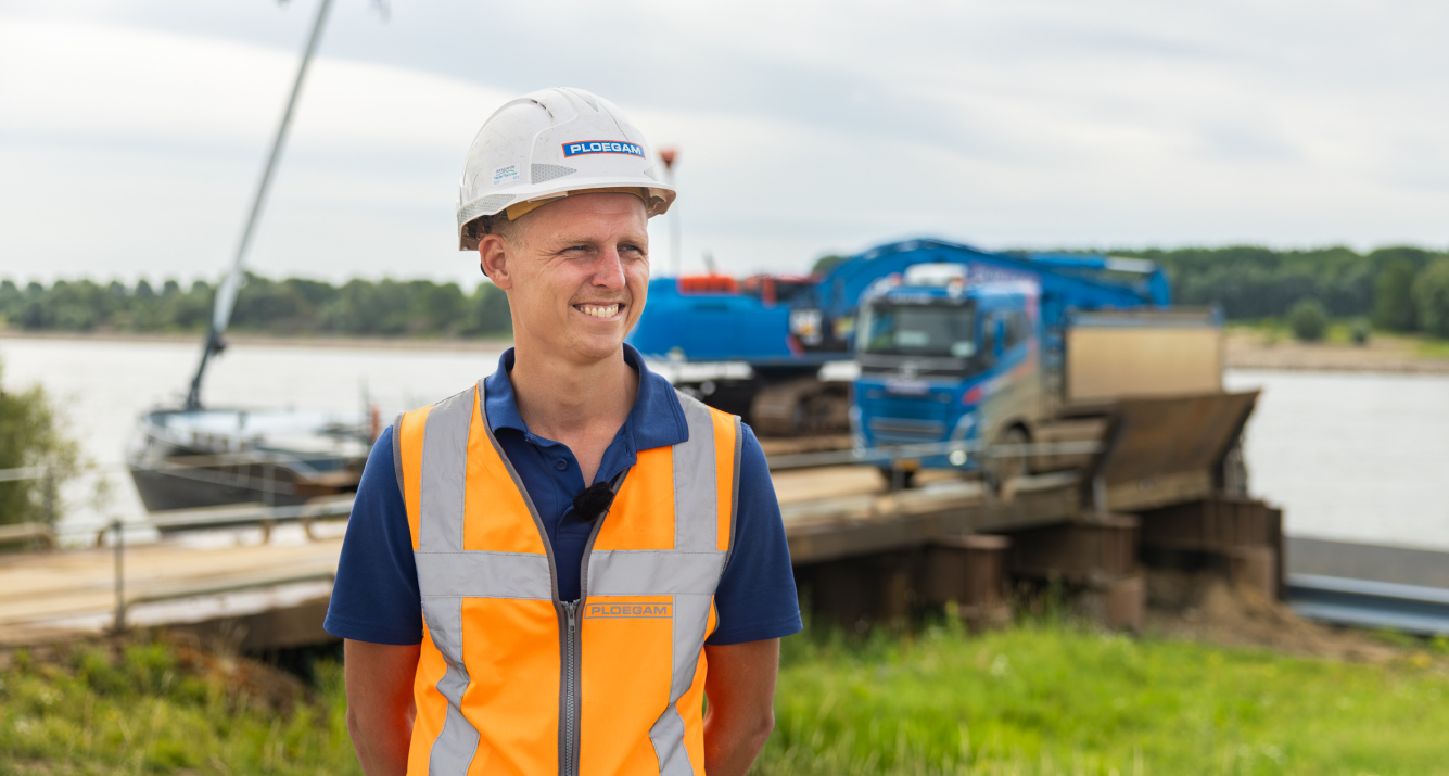 Man met oranje jas en witte Ploegam helm met een blauwe kraan op de achtergrond