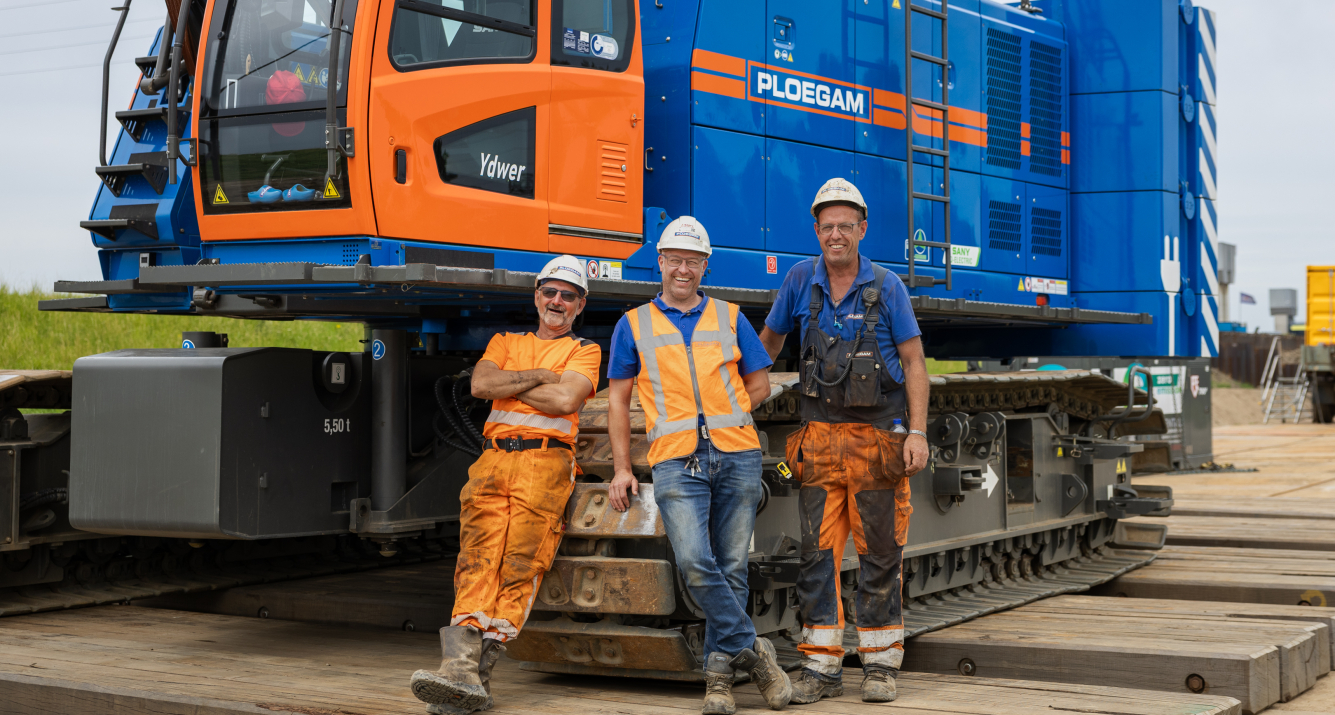 Man met oranje jas en witte Ploegam helm met een blauwe kraan op de achtergrond