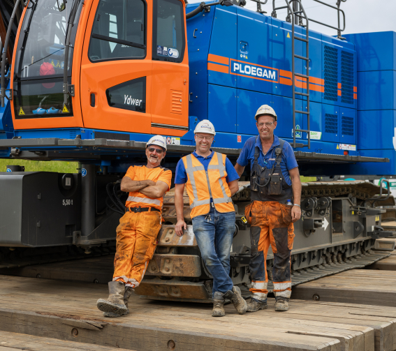 Man met oranje jas en witte Ploegam helm met een blauwe kraan op de achtergrond