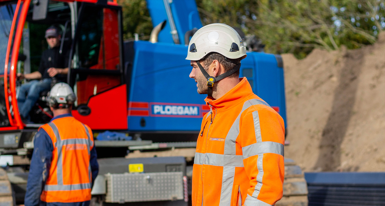 Man met veiligheidsjas en helm op bouwplaats met blauwe kraan op de achtergrond
