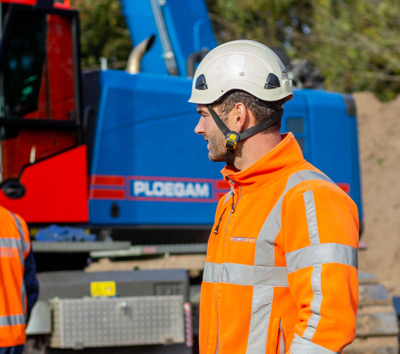 Man met veiligheidsjas en helm op bouwplaats met blauwe kraan op de achtergrond