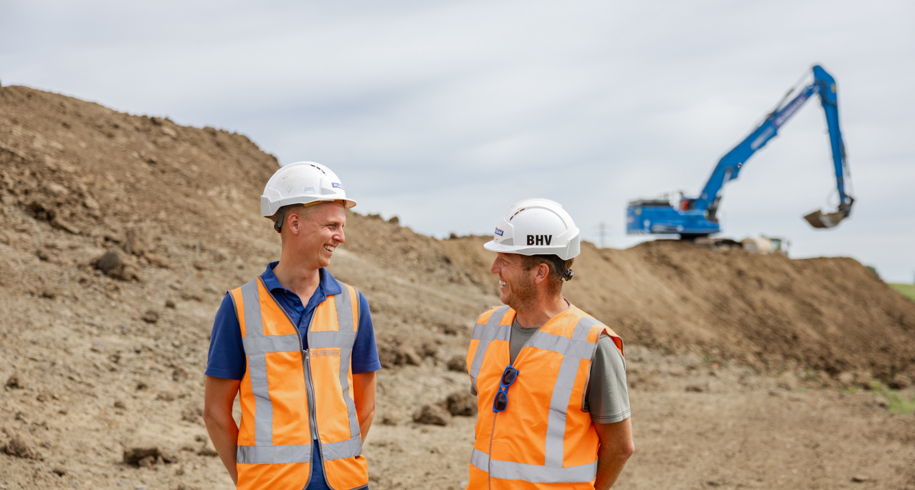 Man met oranje jas en witte Ploegam helm met een blauwe kraan op de achtergrond